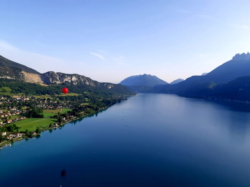 Lac entouré de montagnes verdoyantes avec un ballon rouge en vol, sous un ciel bleu clair, montrant des villages et des champs le long des rives du lac.