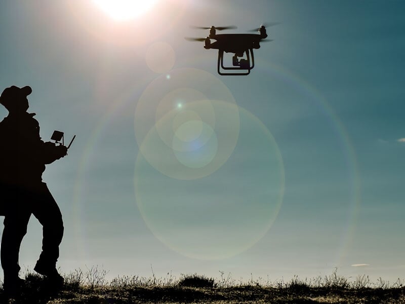 Silhouette d'un homme pilotant un drone contre-jour, ciel bleu avec éclats solaires