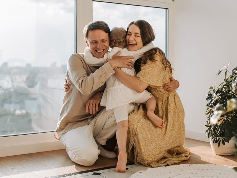 Un couple souriant embrassant leur jeune enfant dans un salon lumineux avec une grande fenêtre et une plante verte.