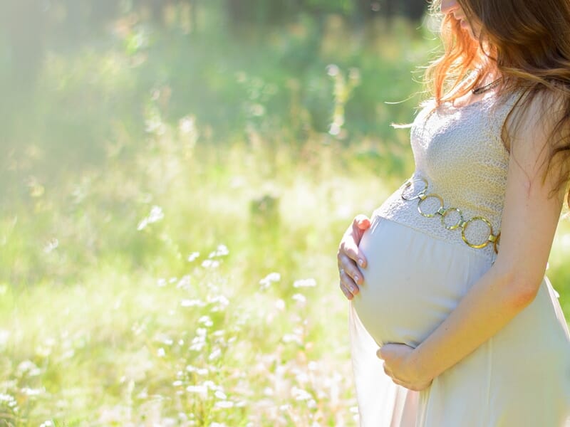 Femme enceinte en robe blanche tenant son ventre dans un champ ensoleillé, entourée de verdure et de lumière douce.