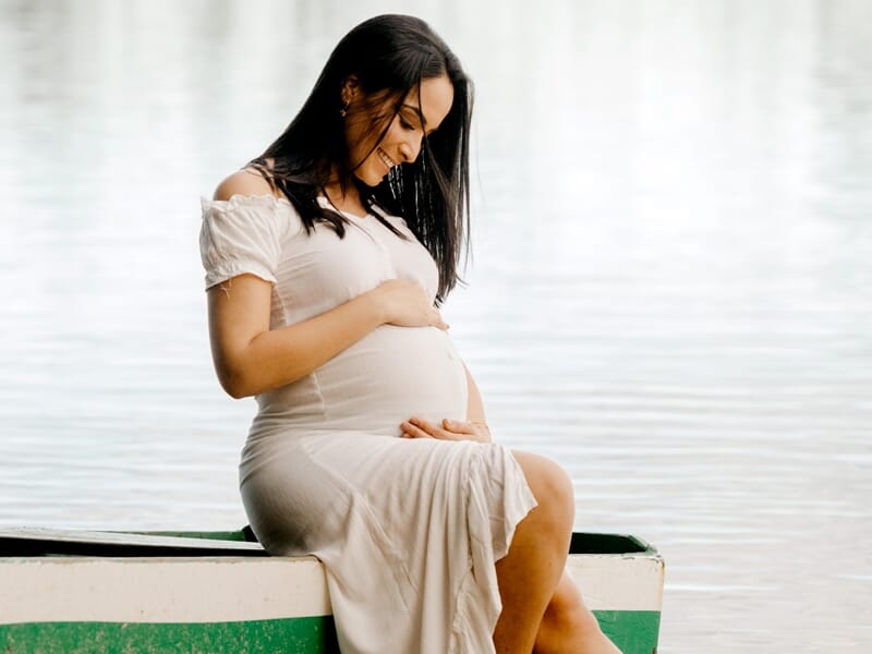 Jeune femme enceinte assise sur un bateau au bord de l'eau, caressant son ventre avec un sourire paisible.