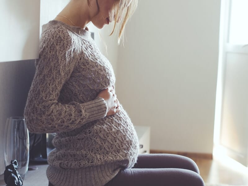Femme enceinte en pull gris assise à l'intérieur, tenant son ventre avec tendresse près d'une fenêtre lumineuse.
