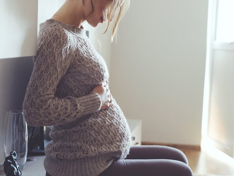 Femme enceinte en pull gris assise à l'intérieur, tenant son ventre avec tendresse près d'une fenêtre lumineuse.