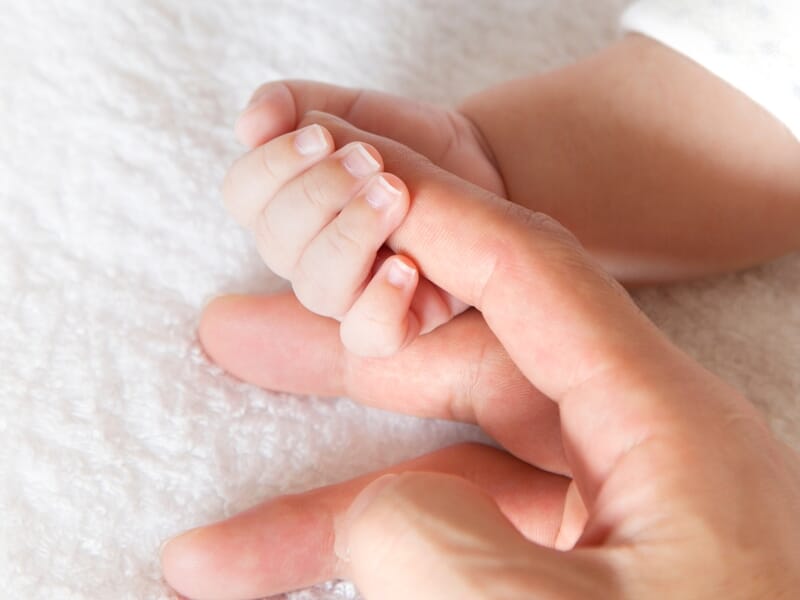 Gros plan sur une main d'adulte tenant délicatement la main d'un bébé sur un tissu blanc doux.