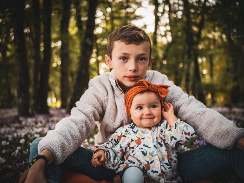 Un garçon assis dans une forêt avec un bébé souriant sur ses genoux, entourés d'arbres et de fleurs.
