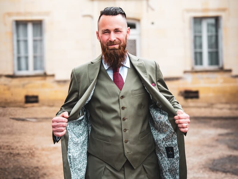 Homme barbu en costume vert ouvrant sa veste pour montrer la doublure, posant devant un bâtiment ancien.