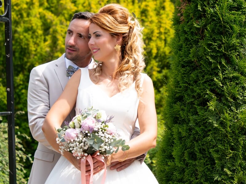 Couple de mariés posant dans un jardin, la mariée tient un bouquet de fleurs, ambiance joyeuse et romantique.