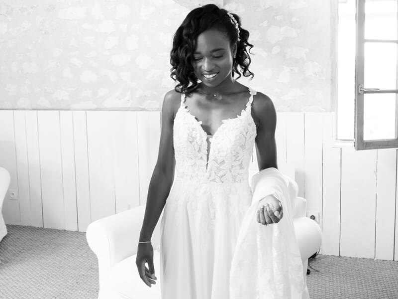Jeune mariée souriante en robe blanche dans une pièce lumineuse, tenant son voile avec élégance.