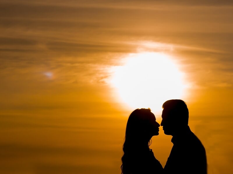 Silhouettes d'un couple face à face au coucher du soleil, ciel orange et ambiance romantique en arrière-plan.