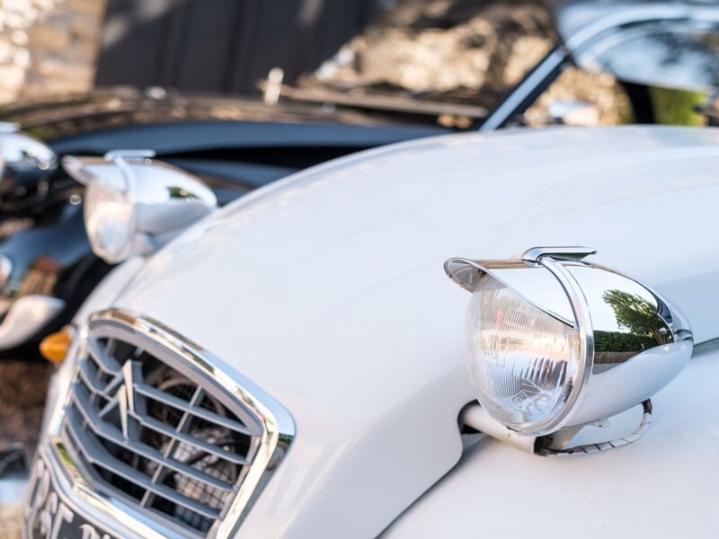 Gros plan sur les phares ronds et chromés de la Citroën 2CV blanche, avec la calandre emblématique ornée du logo Citroën