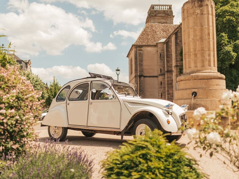 Citroën 2CV blanche garée devant un monument historique en pierre, entourée de végétation, sous un ciel partiellement nuageux