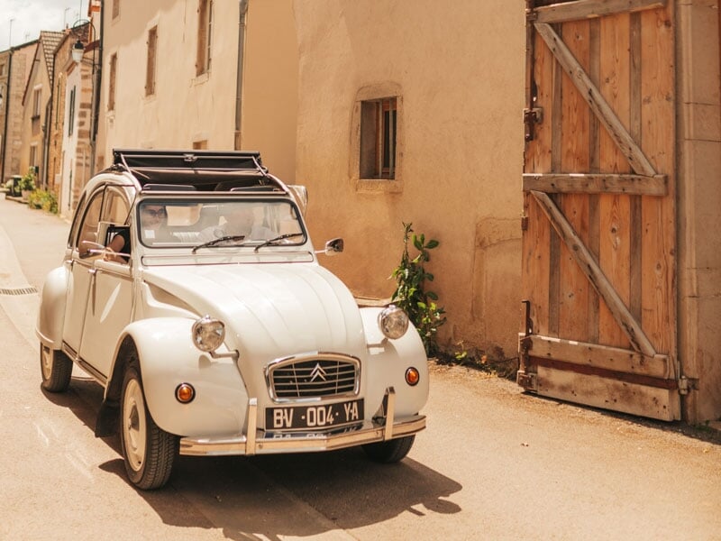 Citroën 2CV blanche circulant dans une rue de village aux façades anciennes, sous un ciel ensoleillé, avec la capote ouverte