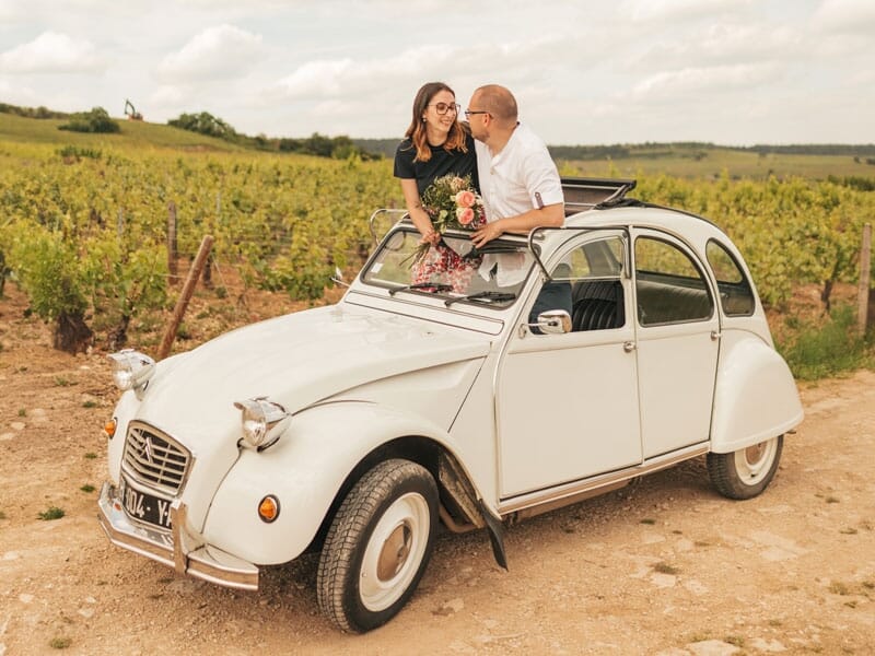 Couple souriant, debout dans une Citroën 2CV blanche décapotée, avec vue sur des vignes en arrière-plan