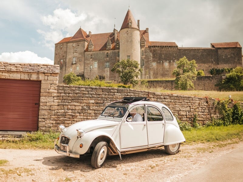 Citroën 2CV blanche devant un château médiéval en pierre, sous un ciel nuageux, avec des remparts en arrière-plan