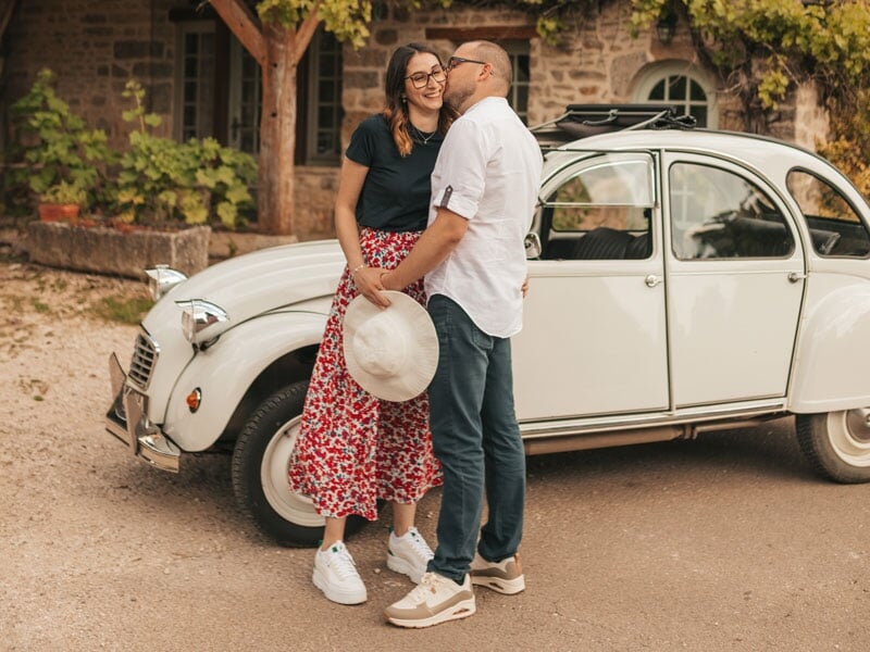 Couple souriant échangeant un regard complice devant une Citroën 2CV blanche, près d’une maison en pierre entourée de végétation