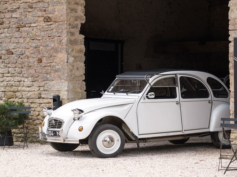Voiture Citroën 2CV blanche garée devant un bâtiment en pierre avec toit ouvrant noir, près de deux chaises en métal noir