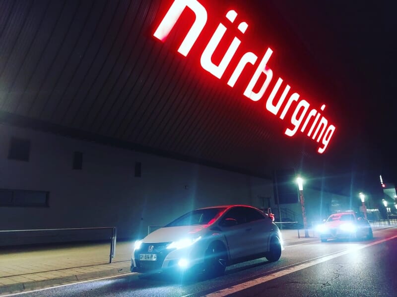 Honda Civic Type R stationnée devant l'entrée illuminée du Nürburgring la nuit, avec une enseigne rouge éclairée, créant un contraste dramatique et mettant en avant l'aspect sportif du véhicule.