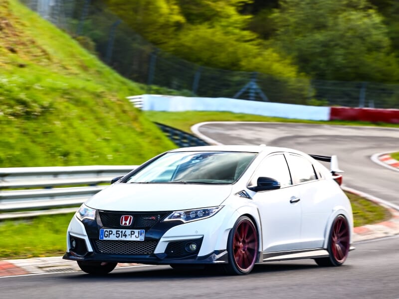 Honda Civic Type R blanche, vue de trois quarts avant en mouvement rapide sur un circuit vallonné avec des barrières de sécurité, entourée d'arbres verts.