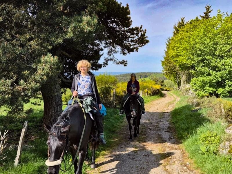 Deux cavalières souriantes sur un sentier de terre bordé d'arbres et de végétation verte.