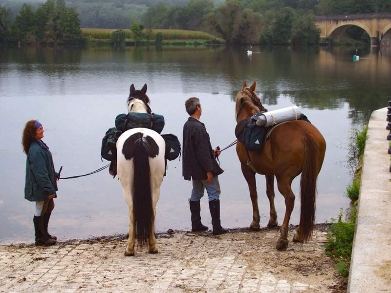 Deux personnes tenant des chevaux chargés de sacs, au bord d'une rivière avec un pont en arrière-plan.