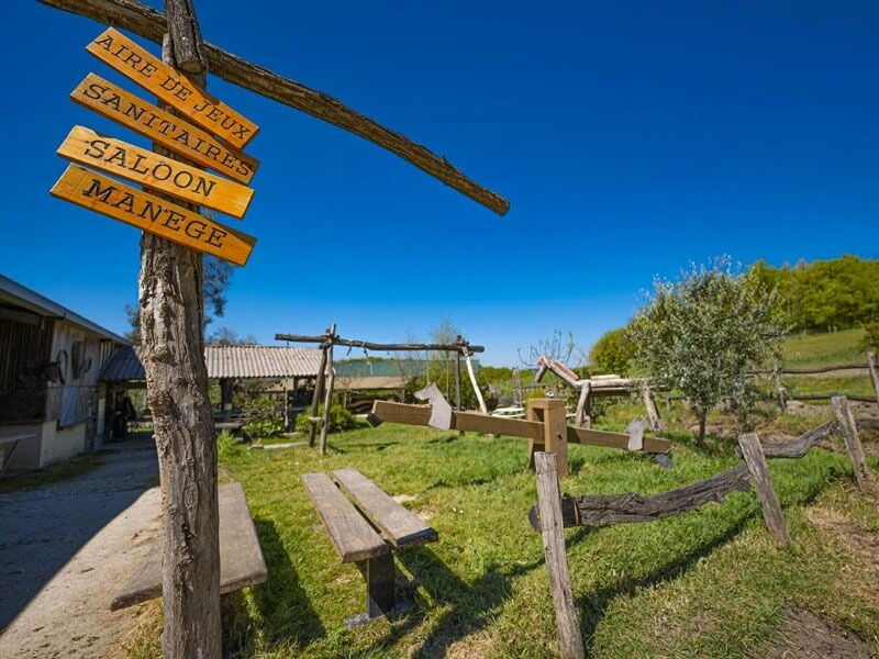 Panneaux en bois indiquant aire de jeux, sanitaires, saloon et manège dans un espace vert avec bancs et balançoires.