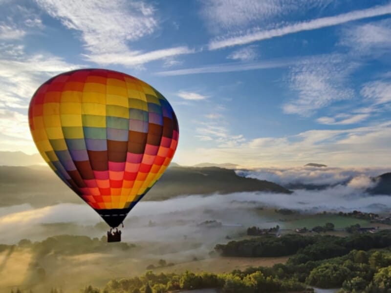 Vol d'un ballon à air chaud multicolore au-dessus d'une vallée enveloppée de brume matinale, avec des montagnes à l'horizon sous un ciel nuageux.