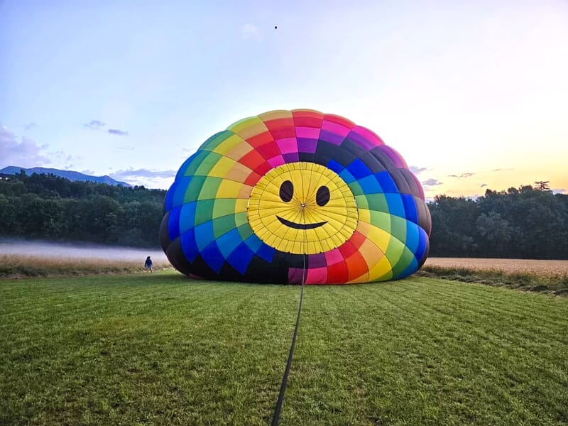 Montgolfière colorée avec un visage souriant déployée sur un champ vert à l'aérodrome de Gap-Tallard au lever du jour.
