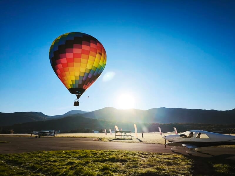 Montgolfière colorée en vol au-dessus de l'aérodrome de Gap-Tallard avec un petit avion au sol et le soleil bas.