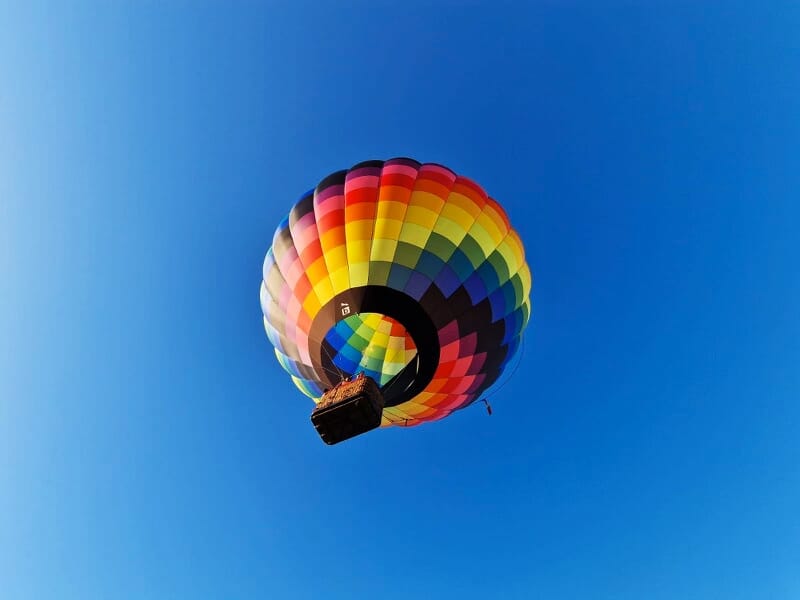Montgolfière multicolore en vol dans un ciel bleu clair, vue en contre-plongée avec la nacelle suspendue.
