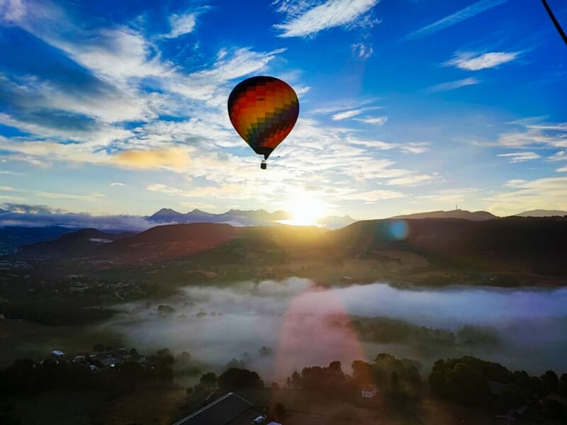 Montgolfière colorée volant au-dessus d'un paysage vallonné avec des nuages bas et un ciel partiellement nuageux.