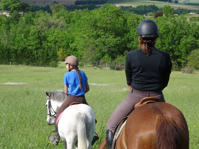 Vue arrière de deux cavalières, la première sur un cheval blanc et la seconde sur un cheval brun, portant des casques de protection et observant un paysage boisé.