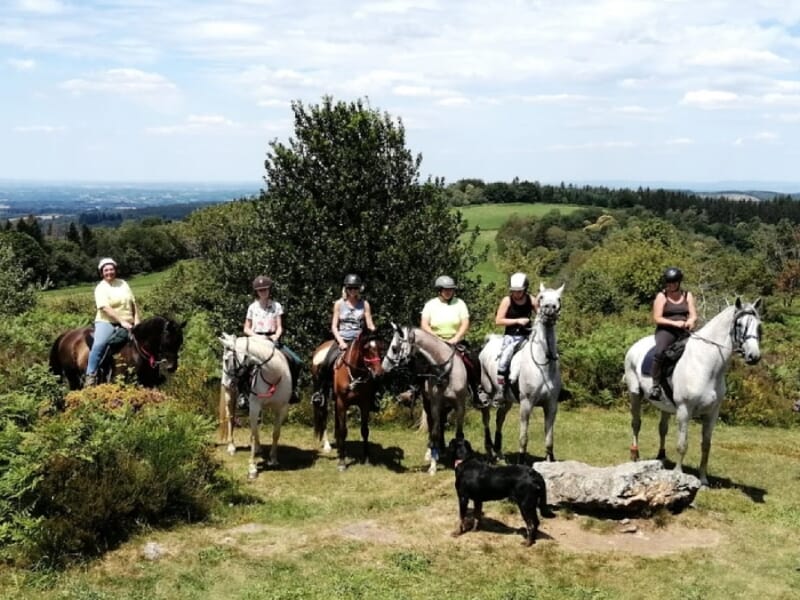 Groupe de cavaliers et de chevaux sur un point de vue élevé, paysage vallonné en arrière-plan, ciel bleu parsemé de nuages, accompagnés d'un chien noir.