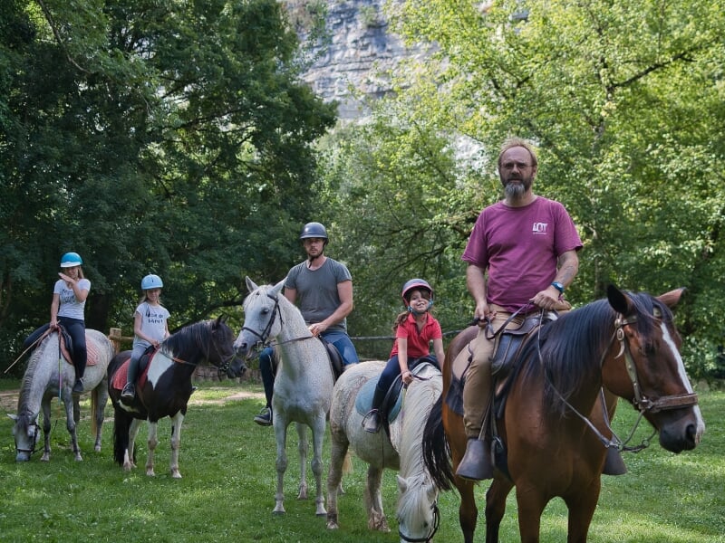 Groupe familial de cinq personnes, incluant un enfant, en balade à cheval dans un parc luxuriant. Le cavalier le plus âgé, en tête, dirige le groupe avec assurance sur son grand cheval brun, tandis qu