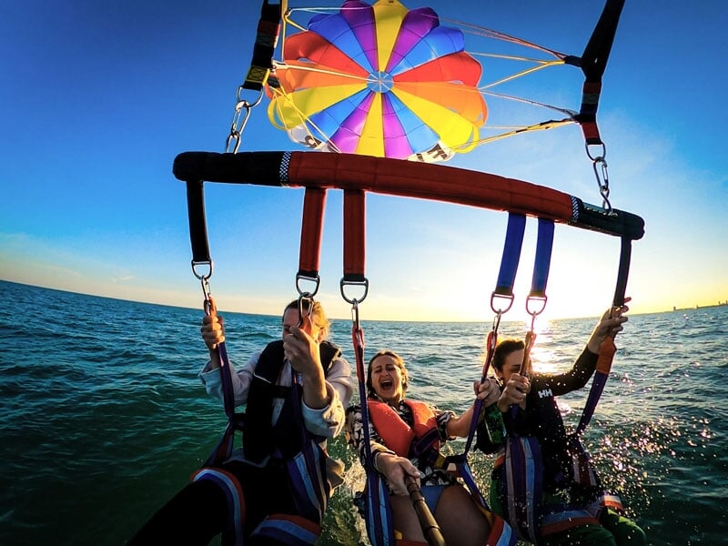 Trois personnes faisant du parachute ascensionnel au-dessus de l'océan au coucher du soleil. Elles sont harnachées et assises, avec un parachute coloré au-dessus d'elles