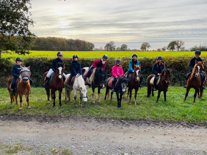 Groupe de cavaliers posant sur leurs chevaux sur un chemin rural, avec un champ de colza en fleur en arrière-plan sous un ciel crépusculaire. Les cavaliers, vêtus d'équipements de protection, montrent