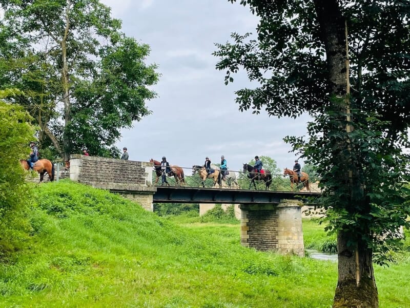 Cavaliers traversant un pont en pierre ancien au-dessus d'un cours d'eau, dans un paysage verdoyant. La scène est encadrée par une végétation luxuriante et une architecture traditionnelle, offrant une