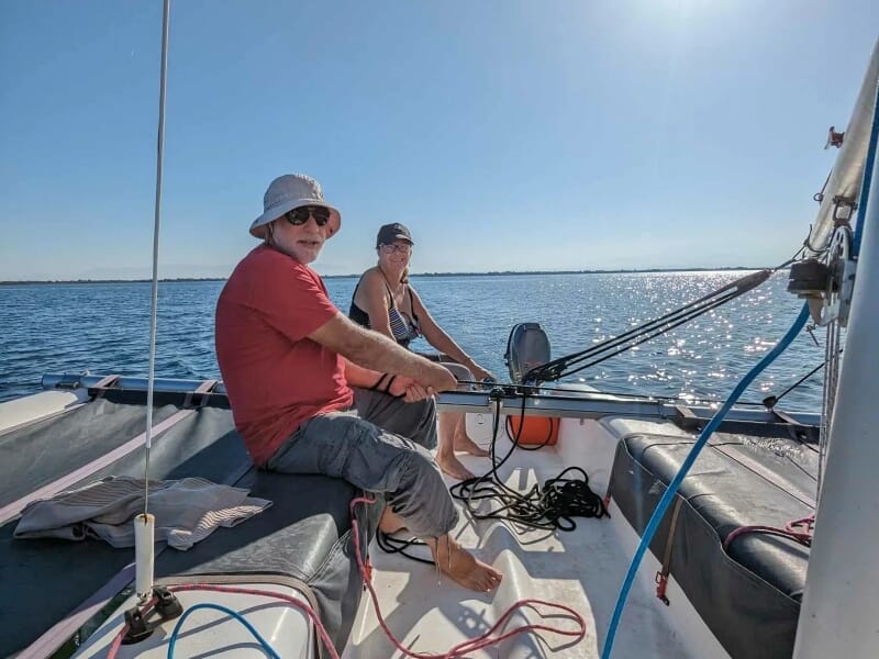 Deux adultes assis à l'arrière d'un voilier, l'homme à la barre portant un t-shirt rouge et un chapeau blanc, et la femme en débardeur bleu, sous un ciel sans nuages avec une étendue d'eau calme en ar