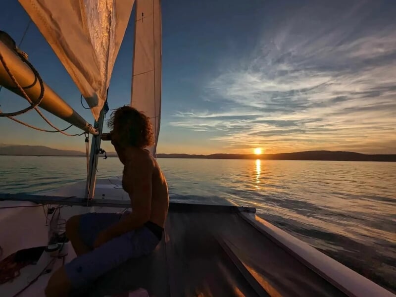 Vue en contre-jour d'un homme assis sur le pont d'un voilier au coucher du soleil, le ciel aux nuances de bleu et d'orange reflétant sur une mer paisible, donnant une sensation de tranquillité et d'is