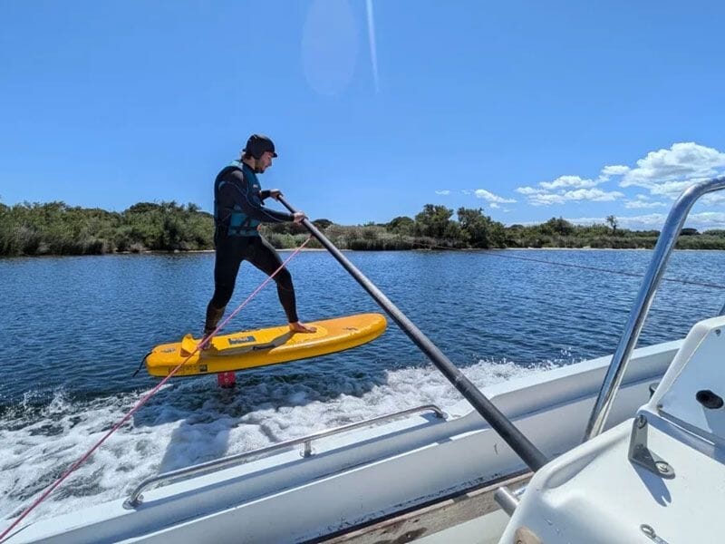 Homme en combinaison debout sur une planche jaune tirée par un bateau sur une eau calme sous un ciel bleu.