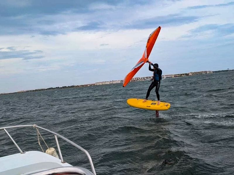 Pratiquant du wingfoil sur une planche jaune avec aile orange, flottant au-dessus de l'eau avec un paysage en arrière-plan.