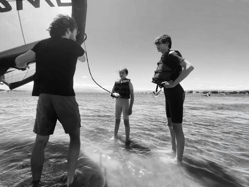 Trois personnes en combinaison discutant sur le rivage, une tenant une aile de wingfoil, photo en noir et blanc.