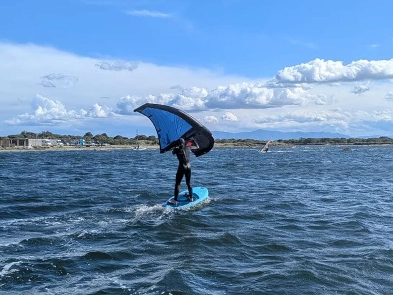 Personne en combinaison sur une planche bleue avec une aile bleue naviguant sur une mer agitée sous un ciel nuageux.
