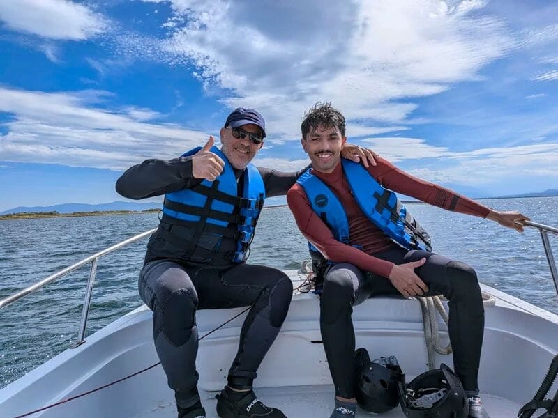 Deux hommes en combinaison et gilet de sauvetage souriants assis à l'avant d'un bateau sur l'eau calme.