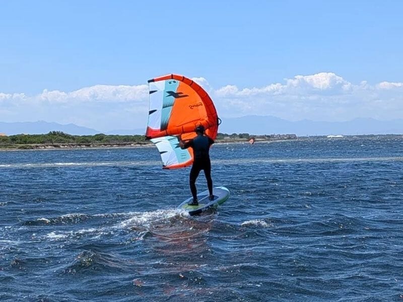 Personne debout sur une planche verte avec une aile orange et blanche, naviguant sur une mer agitée sous un ciel bleu.