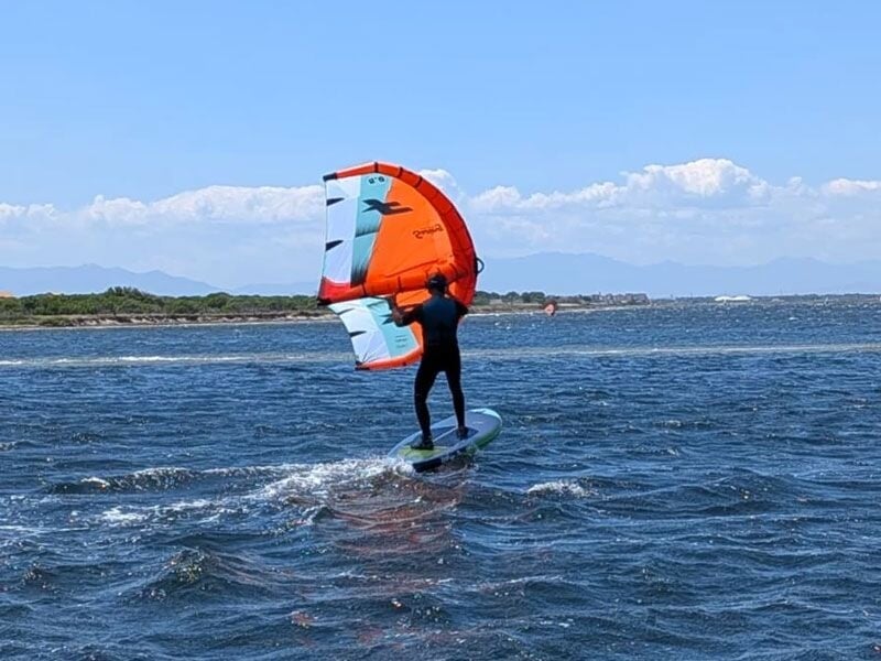 Personne debout sur une planche verte avec une aile orange et blanche, naviguant sur une mer agitée sous un ciel bleu.