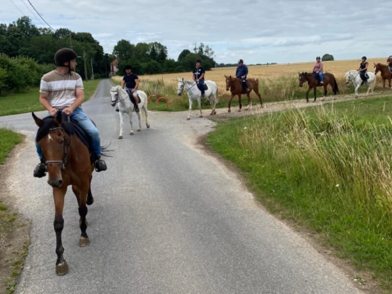 Une rangée de cavaliers parcourt une route campagnarde, entourée de champs verdoyants et d'arbres sporadiques. Un homme en tee-shirt blanc et chapeau mène le groupe sur un cheval brun. Le chemin, fréq