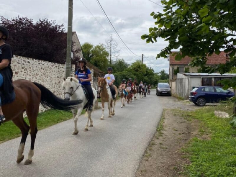 Groupe de cavaliers avançant sur une route de village bordée de maisons en pierre et de verdure. En tête, un cheval blanc est monté par une cavalière en tenue de sport, suivi par un alignement de chev