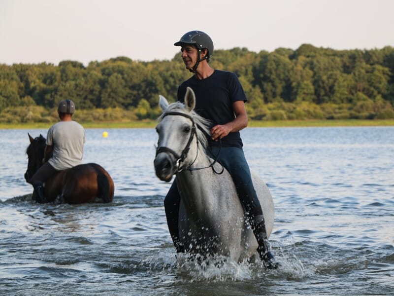 Homme souriant à cheval traversant une rivière, éclaboussant l'eau claire sous un ciel bleu.