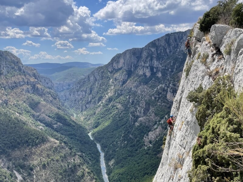 Deux grimpeurs en pleine ascension sur une paroi escarpée des Gorges du Verdon, vallée verdoyante et montagnes en arrière-plan, ciel nuageux.