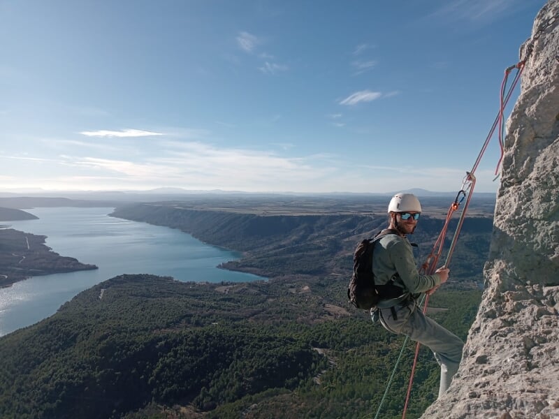 Grimpeur équipé, accroché à une paroi rocheuse avec vue panoramique sur un lac et une vallée boisée, ciel dégagé.
